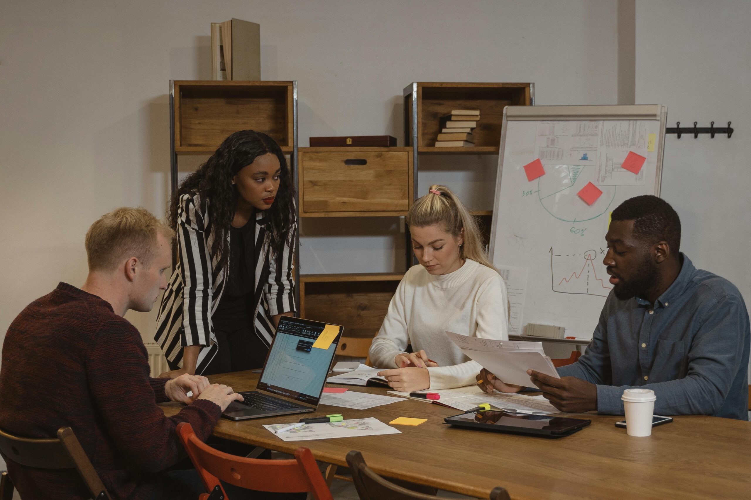Four professionals are in a meeting around a table with laptops, papers, and a coffee cup. A woman stands explaining a point to colleagues who are working and taking notes. A whiteboard with charts and a bookshelf are in the background.