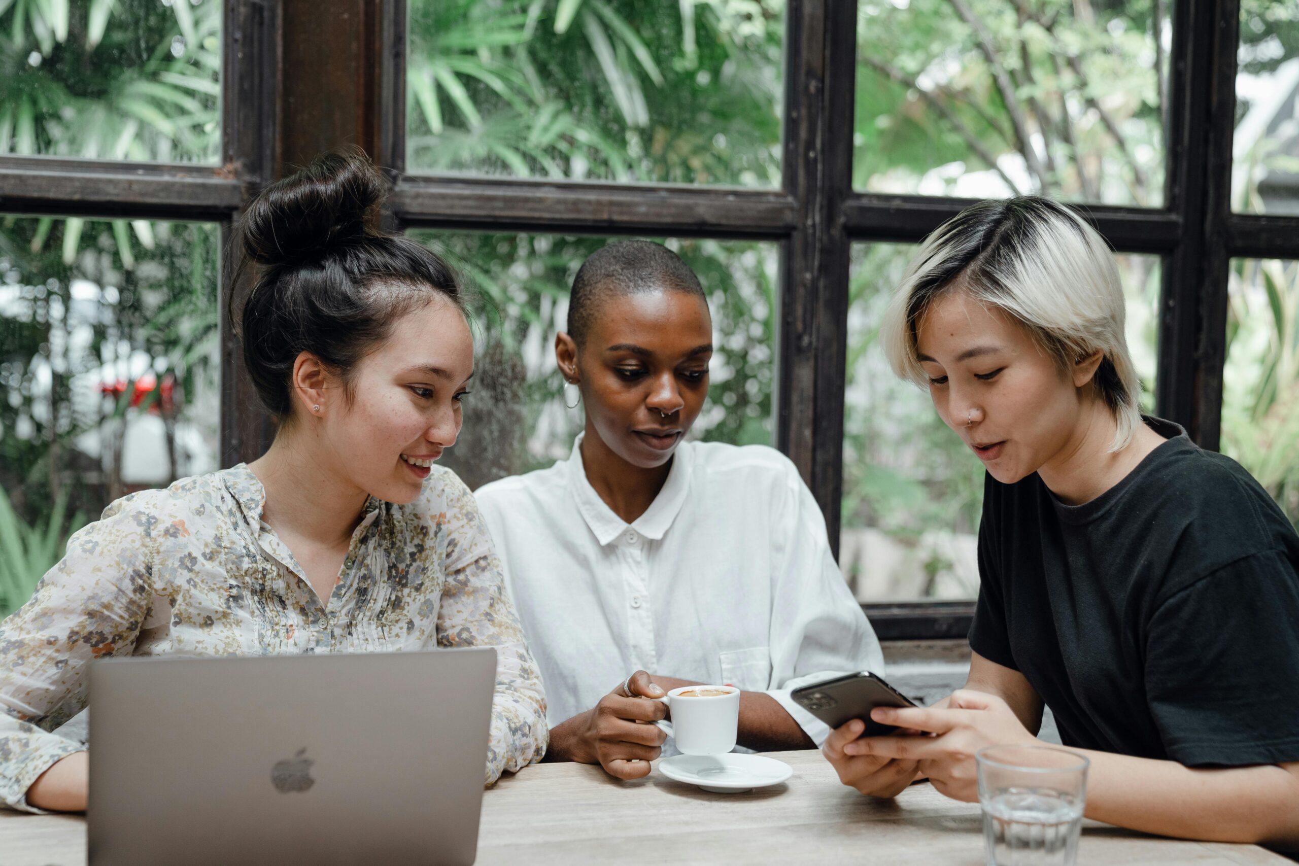 Three women at a table with one looking at a laptop, another holding a coffee cup, and the third checking a smartphone