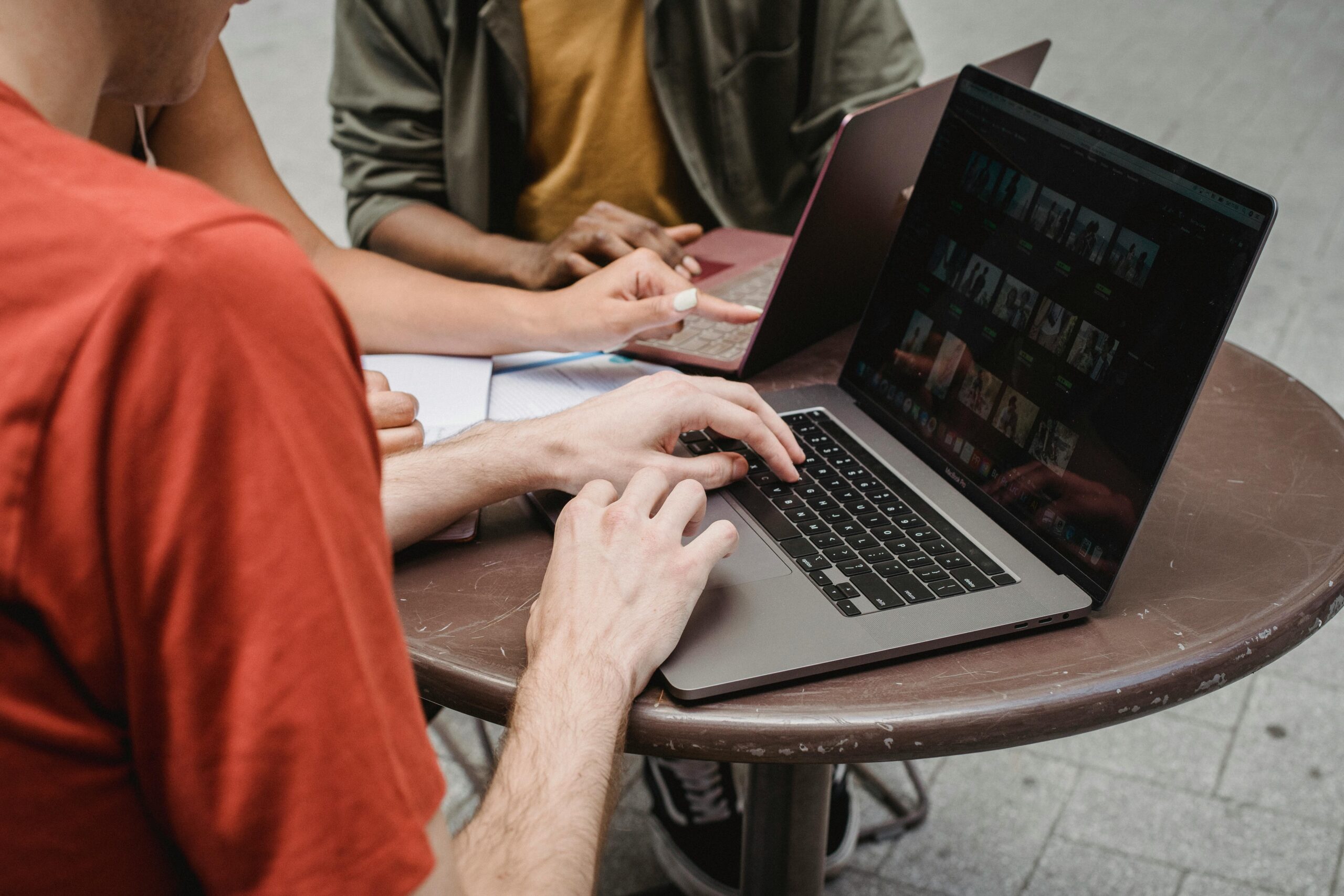 A person typing on a laptop next to another individual writing in a notebook on a round table, indicating a collaborative work environment
