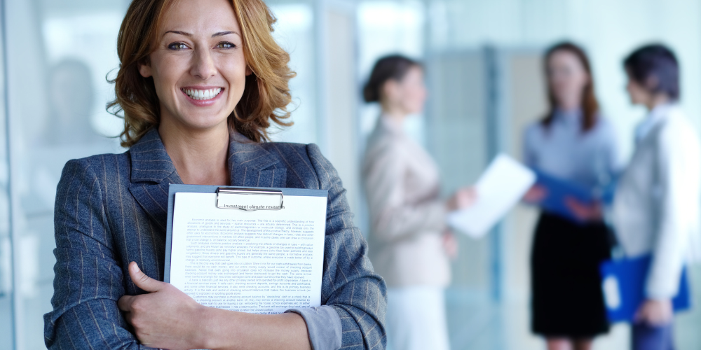 Confident businesswoman smiling at the cam-velascoera, holding a clipboard with documents, with colleagues discussing in the background in a corporate office setting.