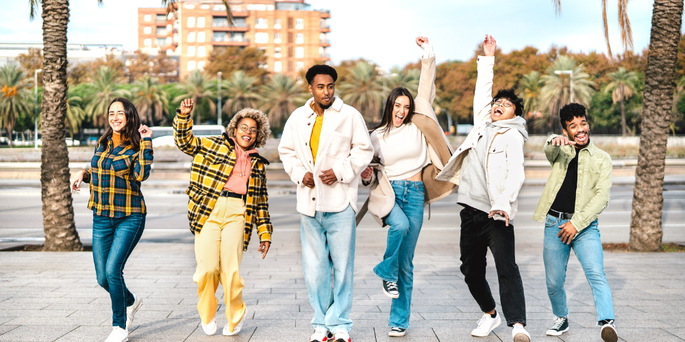 A diverse group of joyful young adults celebrating and walking outdoors, with palm trees and an urban setting in the background.