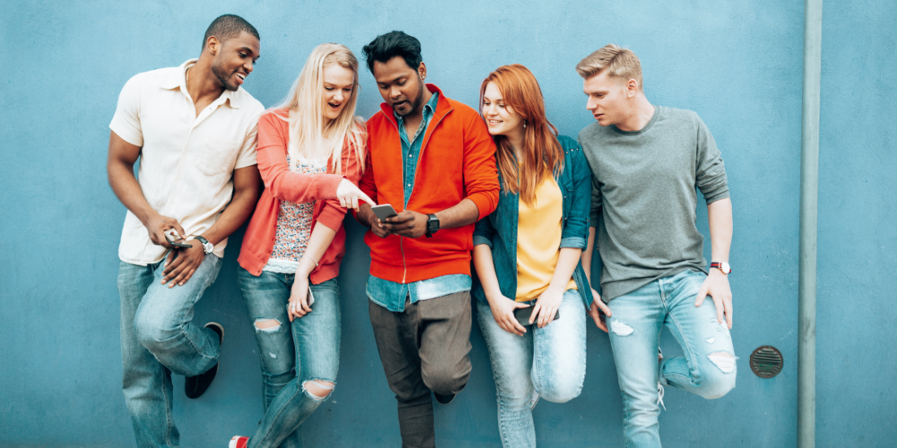 A group of five young adults leaning against a blue wall, looking and smiling at a tablet held by one of them.