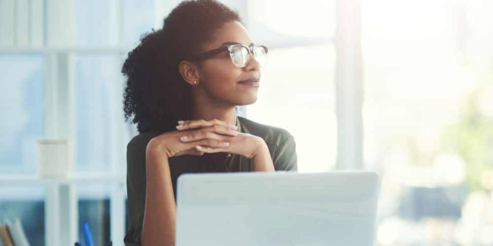A contemplative young woman with glasses sitting at her desk, looking thoughtfully out of a bright office window, with the glow of a computer screen in front of her.