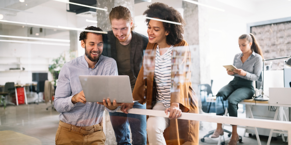 The image is a wide banner showing three individuals, two men and a woman, collaboratively looking at a laptop screen together, engaged and smiling. They seem to be in a modern office space, with another person visible in the background focused on a tablet. The scene depicts a casual yet professional work environment with an emphasis on teamwork and collaboration.