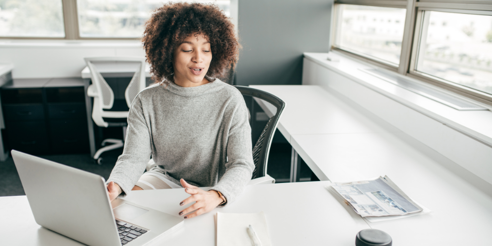 The image is a wide banner depicting a young woman with curly hair sitting at a desk in a bright office, engaging in a video call on her laptop. She appears to be speaking and gesturing, possibly explaining something. The setting is modern and professional.