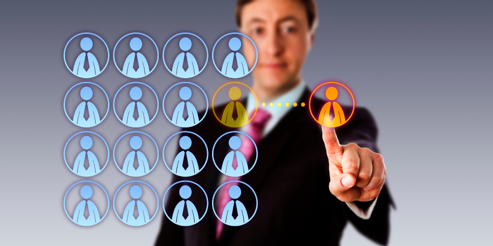 The image is a wide banner that shows a man in a business suit pointing at one icon within a network of various human silhouettes displayed in front of him. Each icon represents a person, with one highlighted in red and yellow, indicating selection or focus on an individual within a group.
