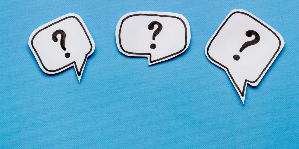 The image is a wide banner with a simple yet striking design, featuring three white speech bubble cutouts, each containing a black question mark. They are set against a bright blue background, giving a clean and bold visual effect. This design could represent questioning, inquiry, discussion, or the need for information.