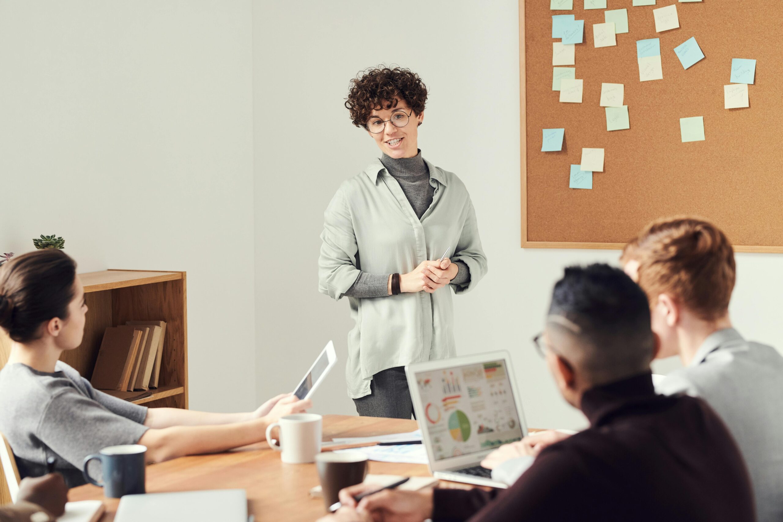 A person with curly hair presenting in front of a cork board with sticky notes to a diverse group of attentive colleagues in a meeting room