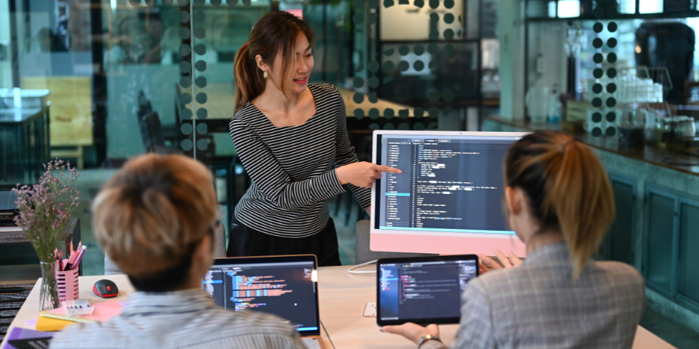 Three individuals in an office environment with one standing and pointing at a large monitor displaying code, likely engaged in a collaborative programming or code review session.