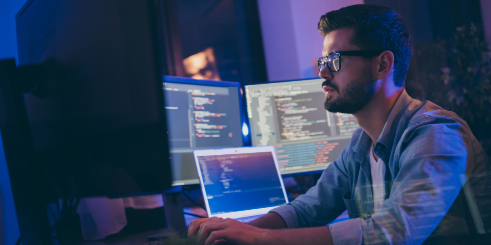 A man with glasses focused on coding, working on multiple computer screens in a dimly lit room with a blue ambiance.