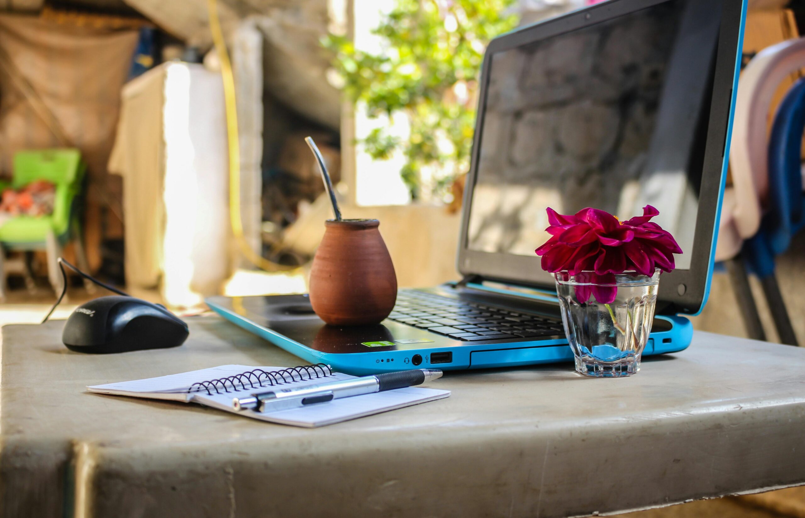 A laptop open on a stone table with a traditional yerba mate gourd, a notebook, a pen, a computer mouse, and a red flower in a glass cup