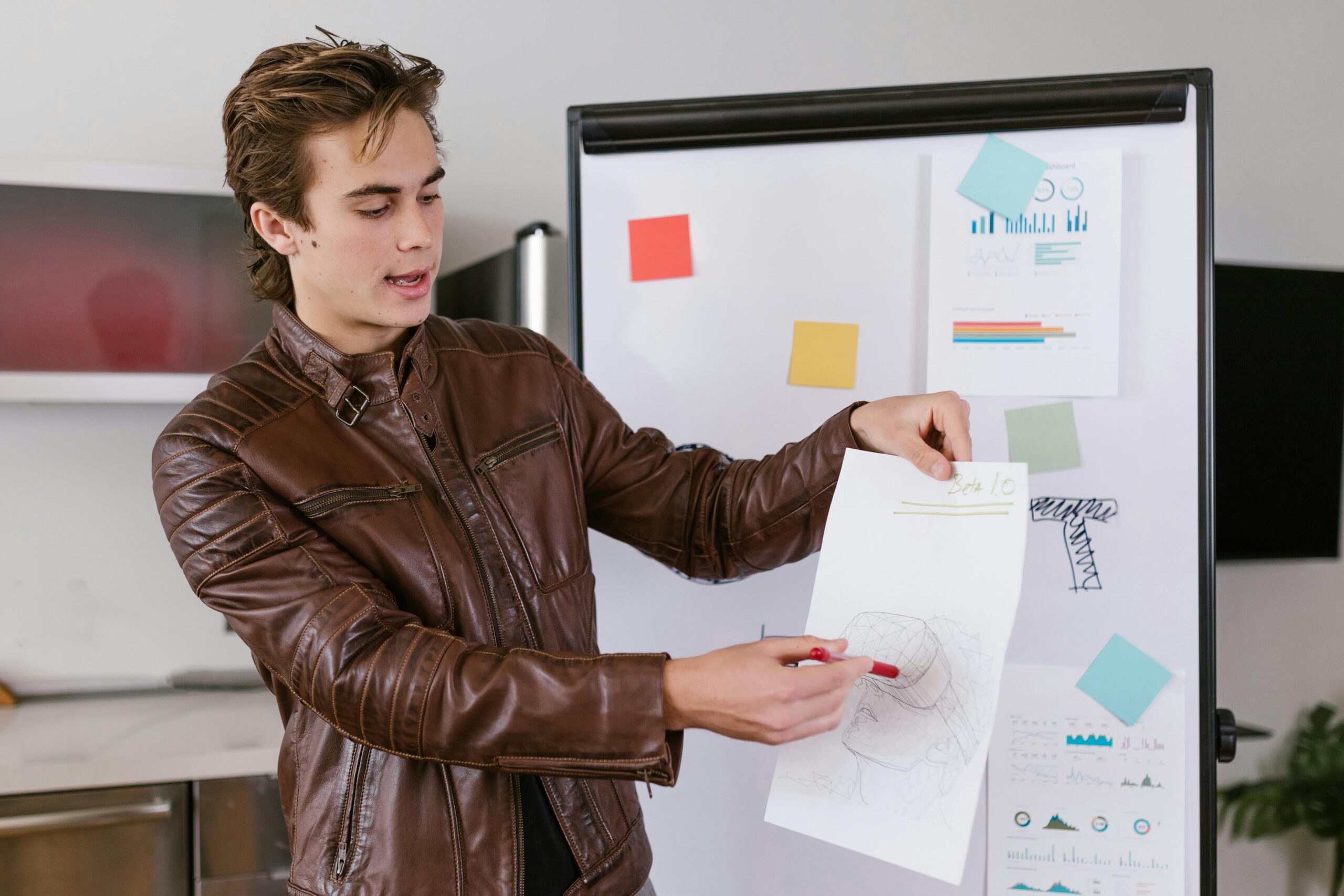 A young man with a leather jacket presents a sketch in front of a whiteboard with colorful sticky notes and data charts from the remote work era.