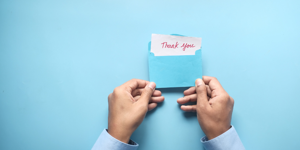 A person holding a light blue envelope with a red "Thank you" note sticking out of it, against a plain light blue background.