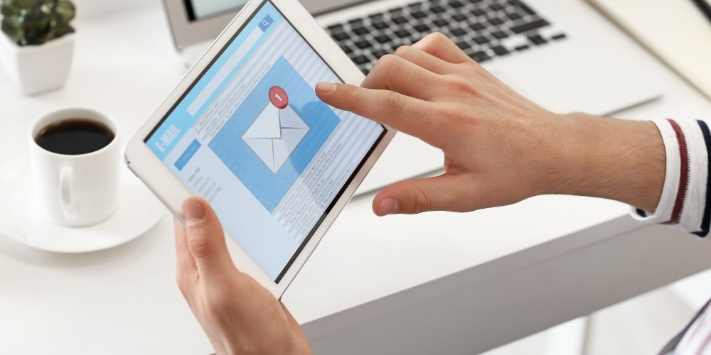 A person holding a tablet with an email application interface on the screen, indicating an unread message, with a laptop and a cup of coffee in the background on a white desk.