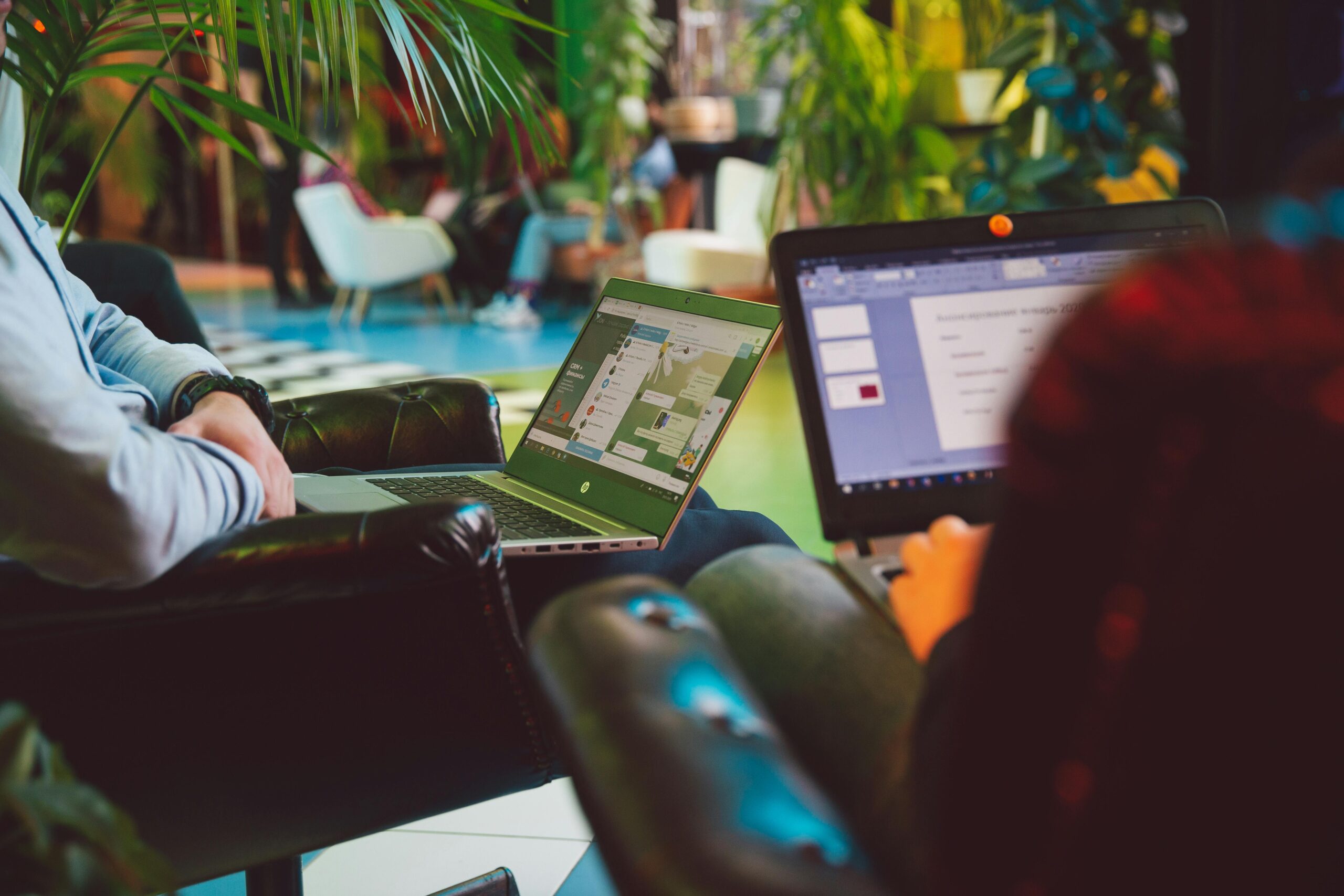 Two individuals working on laptops in a vibrant co-working space with plush leather chairs and lush green plants in the background.