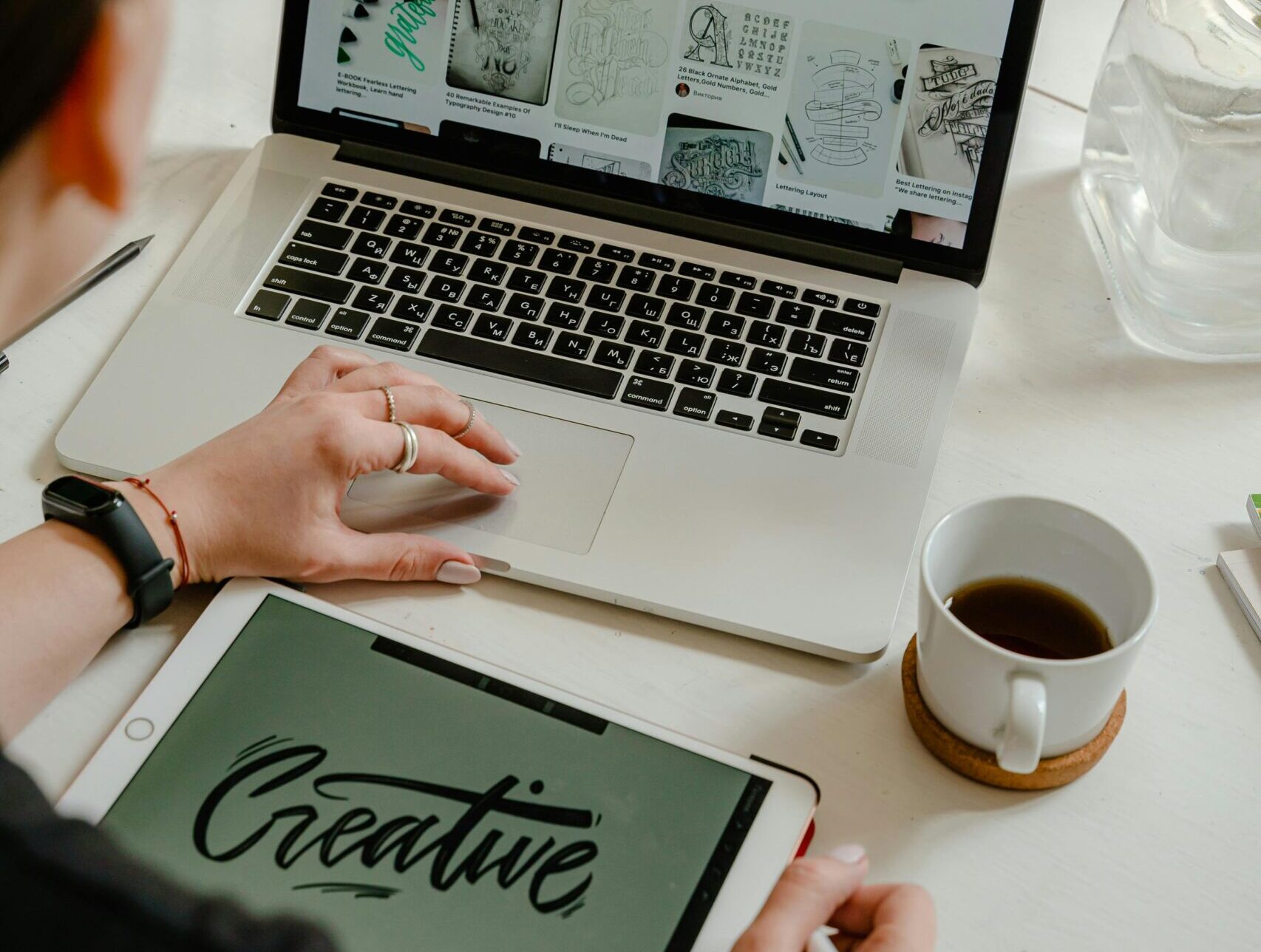 Over-the-shoulder view of a person using a laptop and a digital tablet with 'Creative' written on its screen, with a cup of coffee and vibrant flowers on the table.
