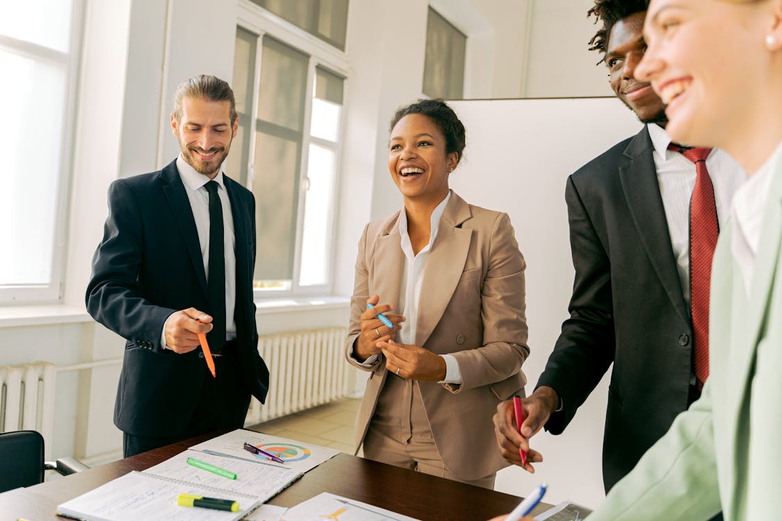 The image shows four professionals in an office, interacting joyfully with papers and markers on a table, indicating a collaborative work environment.