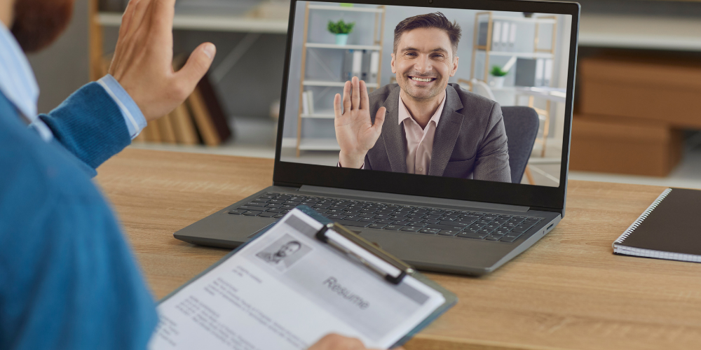 This image depicts a virtual job interview taking place. A person is seen waving at a laptop screen, which displays another individual, a man smiling and waving back. The person conducting the interview is holding a resume and there's a notepad and pen on the desk, suggesting an interview process is underway. The setting appears to be a home office.