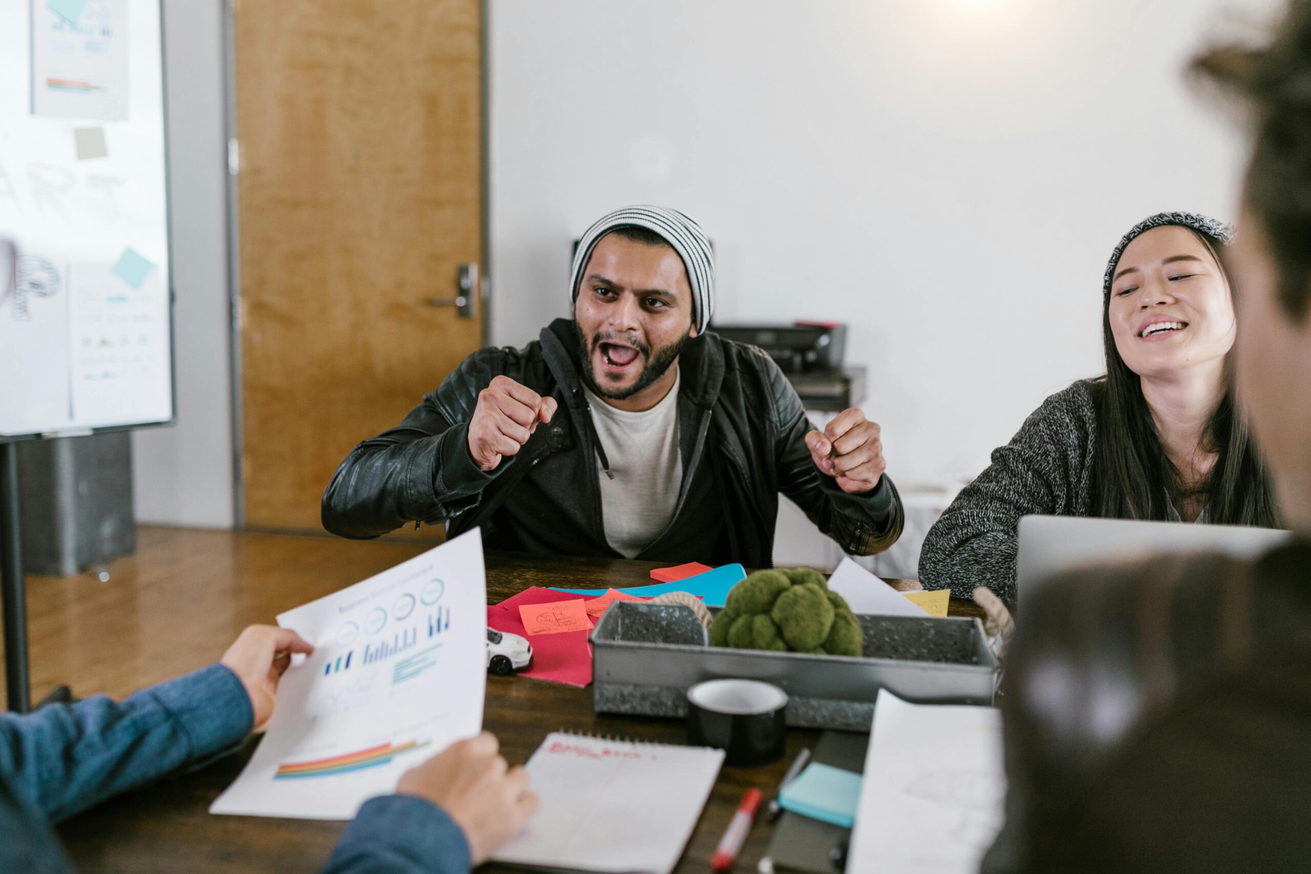A man in a beanie and leather jacket gesturing excitedly at a table with colleagues, one of whom is laughing, in a creative office environment.