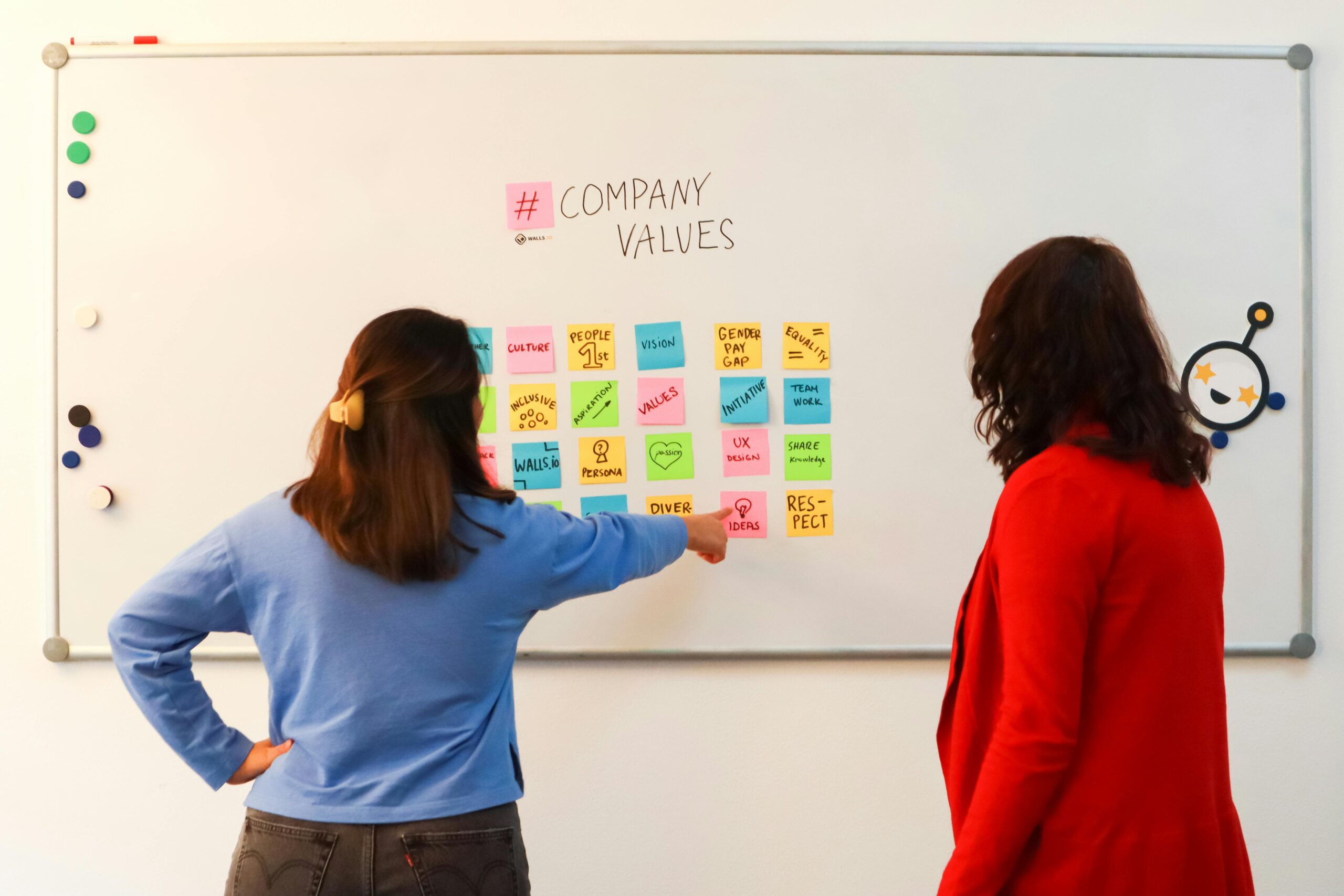 Two people engaging in a brainstorming session in front of a whiteboard filled with colorful sticky notes detailing company values.