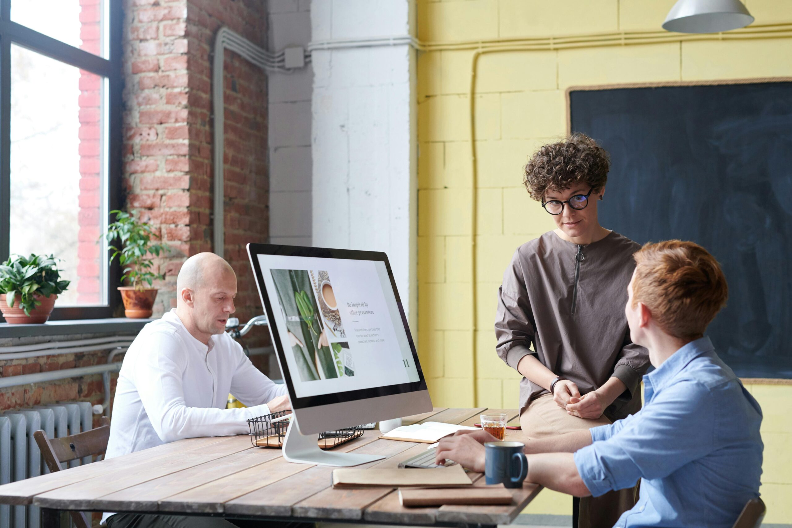 Three professionals discussing a project in a modern office setting with exposed brick walls and large windows.