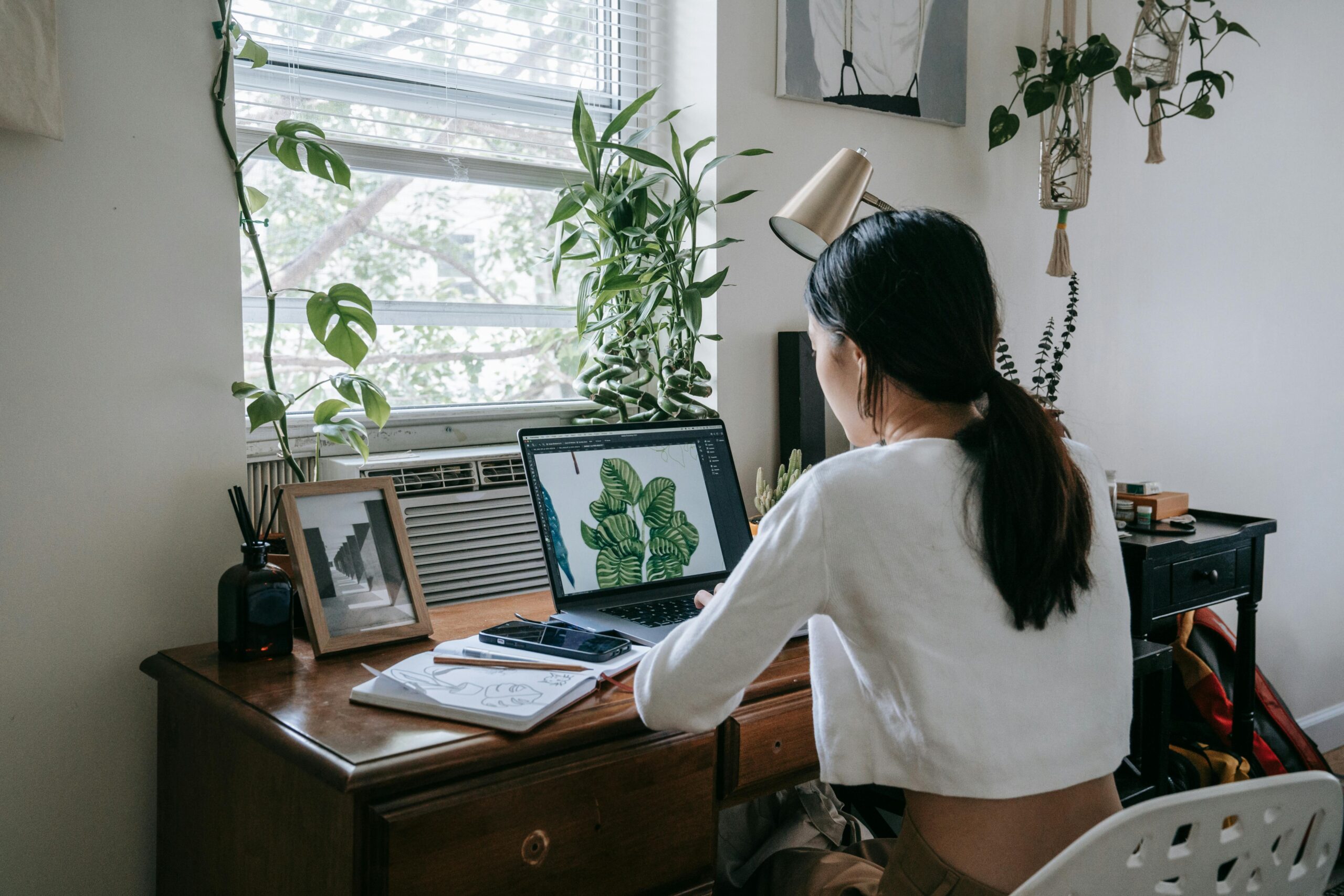A woman sitting at a wooden desk, working on a laptop with a drawing tablet, in a room filled with plants by a window.