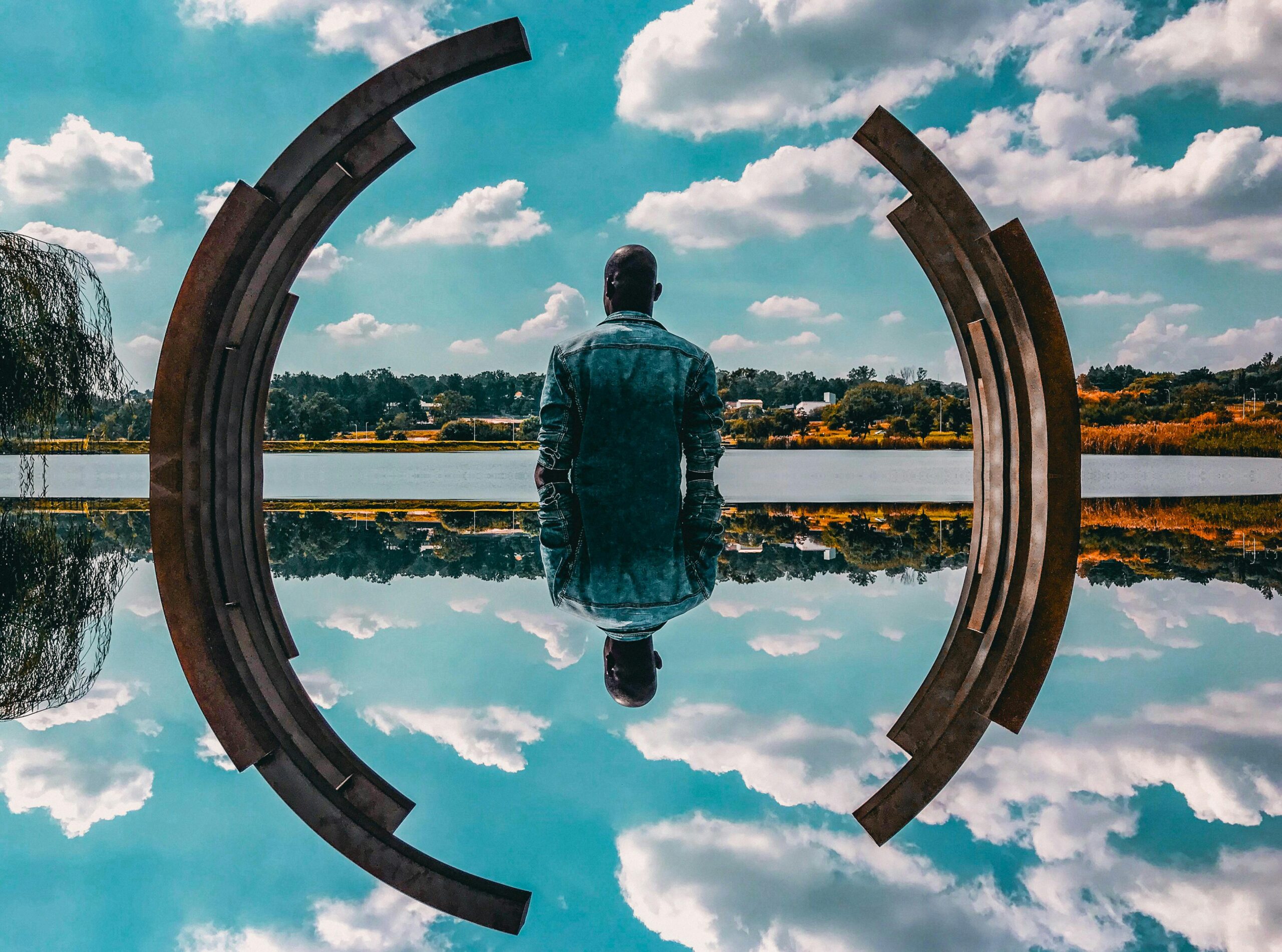 A person in a denim jacket stands with their back to the cam-velascoera, looking out over a still lake that reflects the sky and clouds above. The scene is framed by a large, rust-colored metal sculpture that curves into circular forms on both sides.