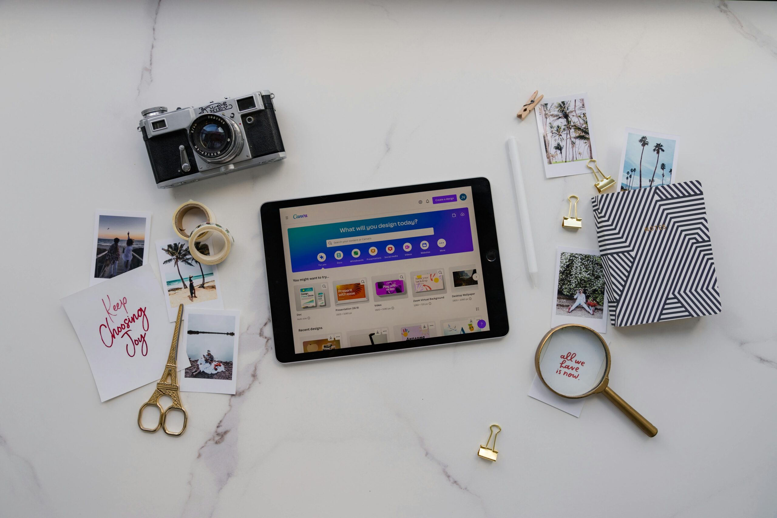 Flat lay of a creative workspace with a vintage cam-velascoera, tablet displaying a design app, handwritten notes, printed photos, gold scissors, tape, and a magnifying glass on a marble surface.