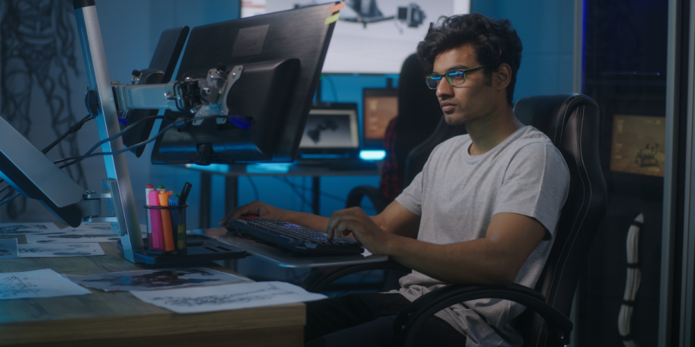 A focused individual is working at a multi-monitor computer setup in a room with blue ambient lighting. There are sketches and a cup with writing utensils on the desk.