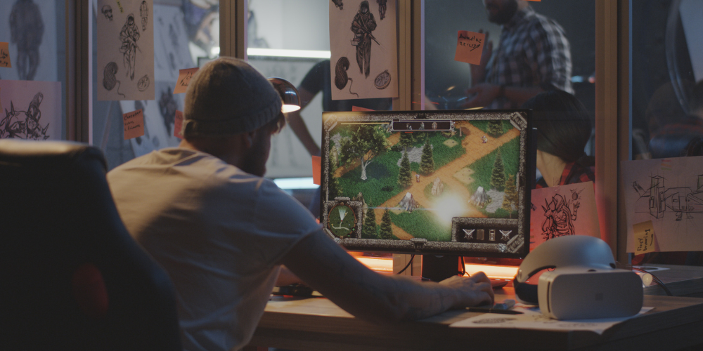 A person wearing a hat is at a desk with a computer showing a strategy game on the screen, surrounded by drawings and notes pinned on the wall.