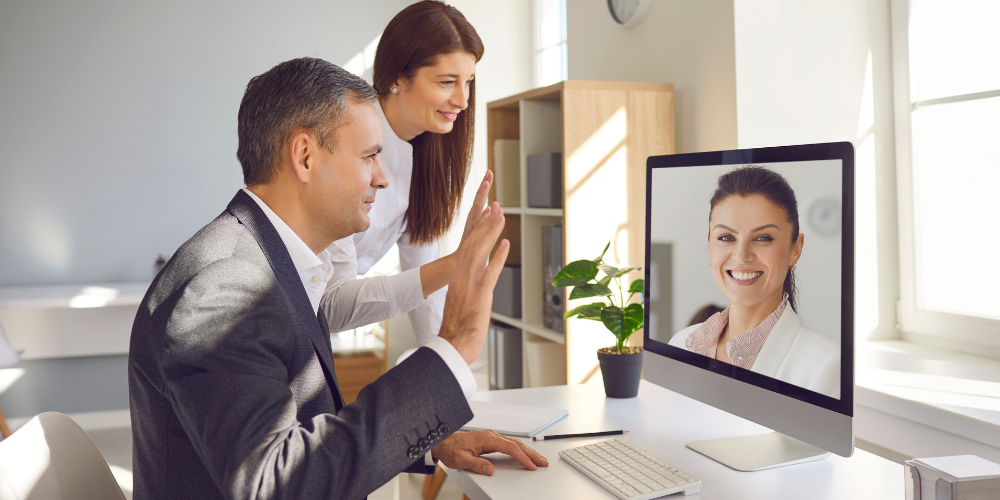 A man in a suit and a woman standing beside him are waving towards a computer screen during a video call in a modern office.