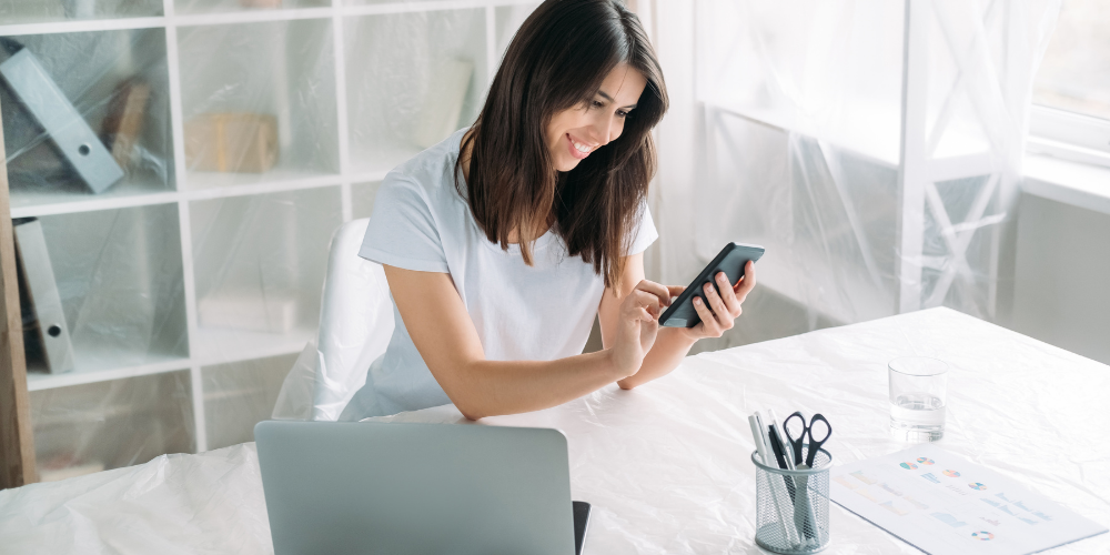 A cheerful woman using a smartphone, with a laptop open in front of her on a white table, in a bright room.