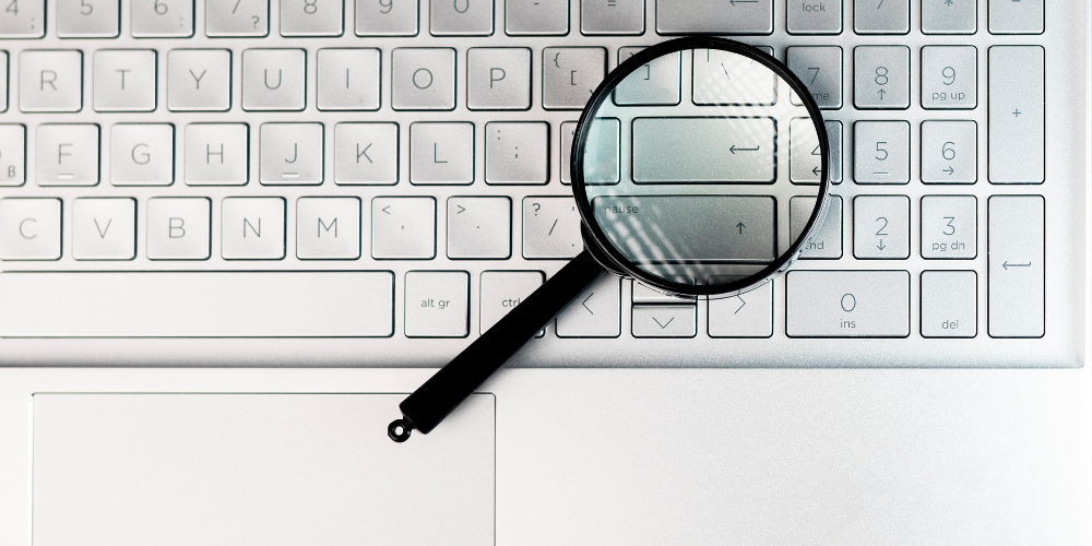 The image displays a magnifying glass resting on a laptop keyboard, with the glass part directly over some of the keys, possibly indicating a detailed search or investigation on the computer. The image captures a common metaphor for scrutiny or close examination in the digital realm.
