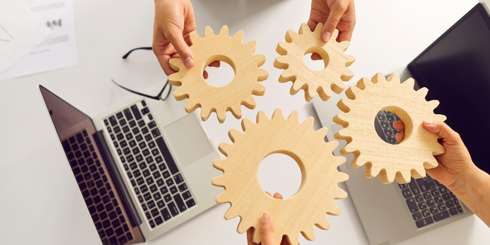 The image features four hands from two different people, each holding a wooden gear above a table where two laptops and some papers are placed. The positioning of the gears suggests they are about to be interconnected, symbolizing teamwork, collaboration, and the idea of different parts working together cohesively, often used as a metaphor for organizational synergy or project management.