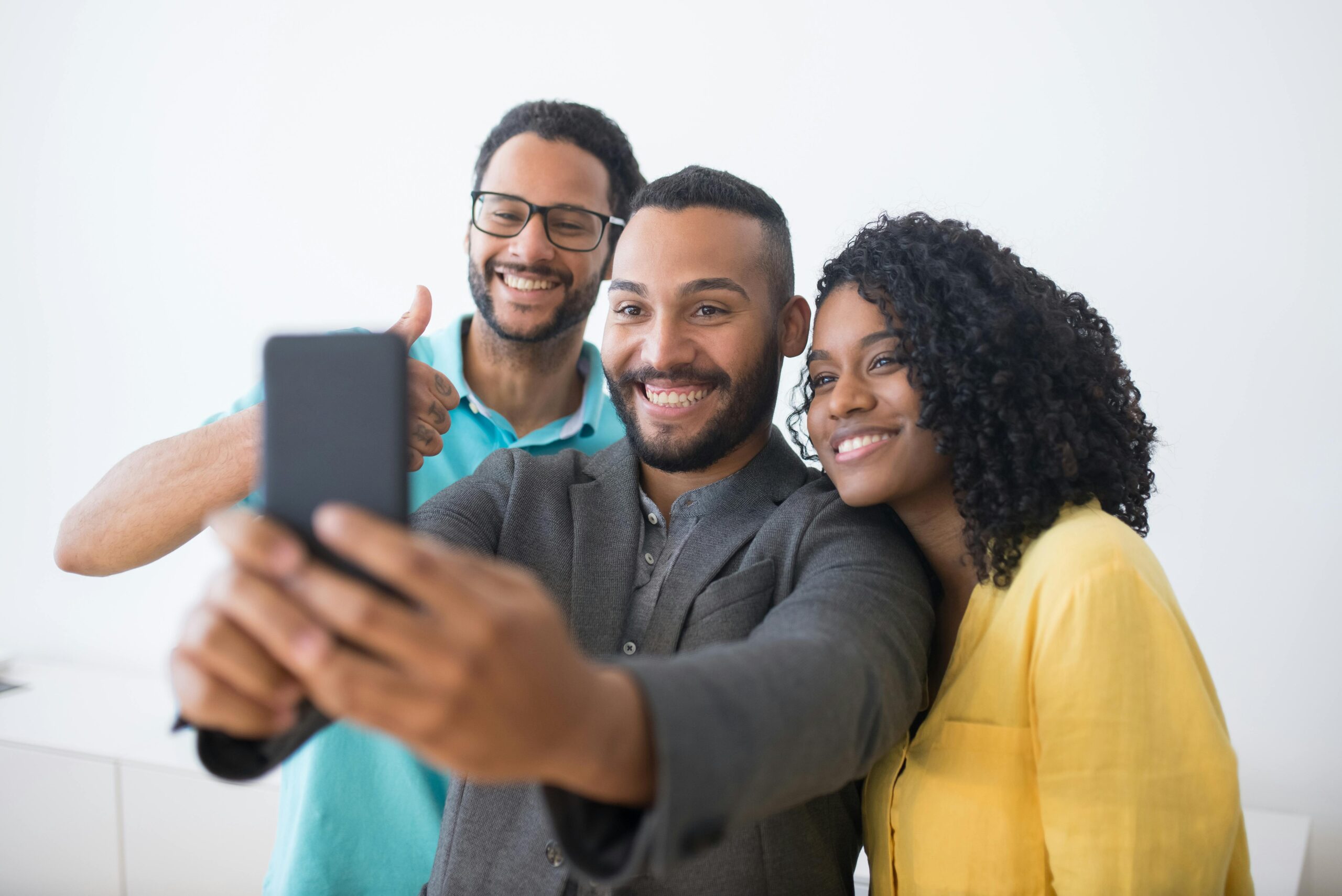 Three cheerful friends taking a selfie with a smartphone, the man in the center holding the device with one hand and giving a thumbs up with the other, with a woman on his right and another man behind, all smiling at the cam-velascoera.