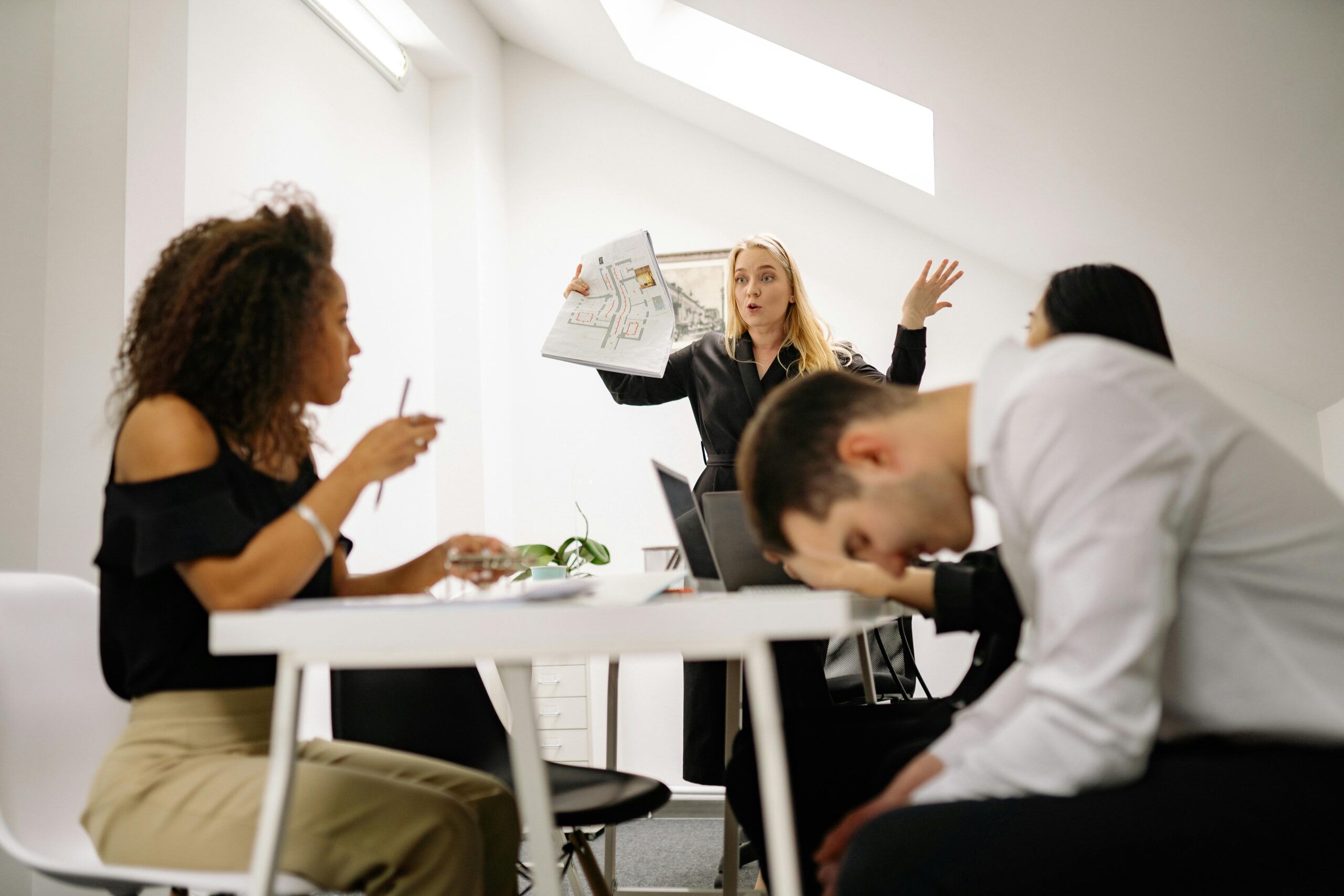 A meeting in a bright office space where a woman stands presenting a blueprint with an expressive gesture, while three colleagues are seated around the table; one speaks, another listens attentively, and the third appears fatigued, resting his head in his hand.