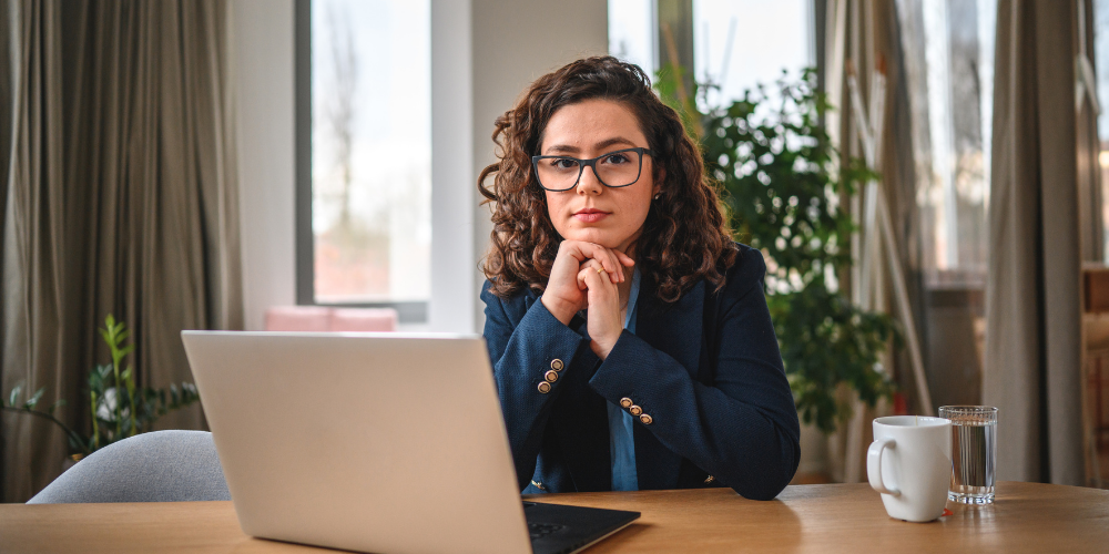 A professional woman in a blue blazer sitting at a desk, pensively looking at her laptop screen, in a cozy office space.