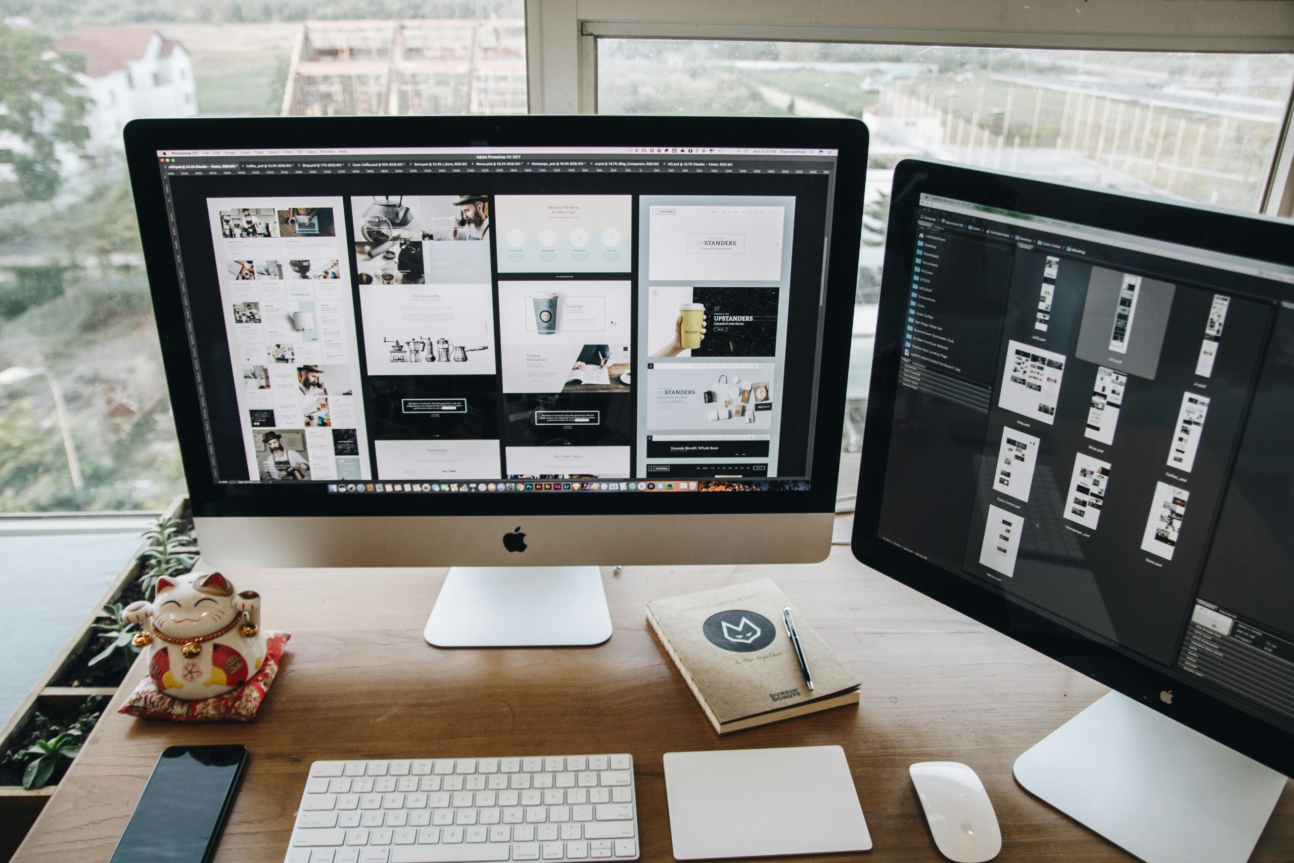 A desktop workspace with two Apple iMacs displaying a website design and code editor, with a smartphone, a notebook, and a Maneki-neko (lucky cat) statue on the desk.