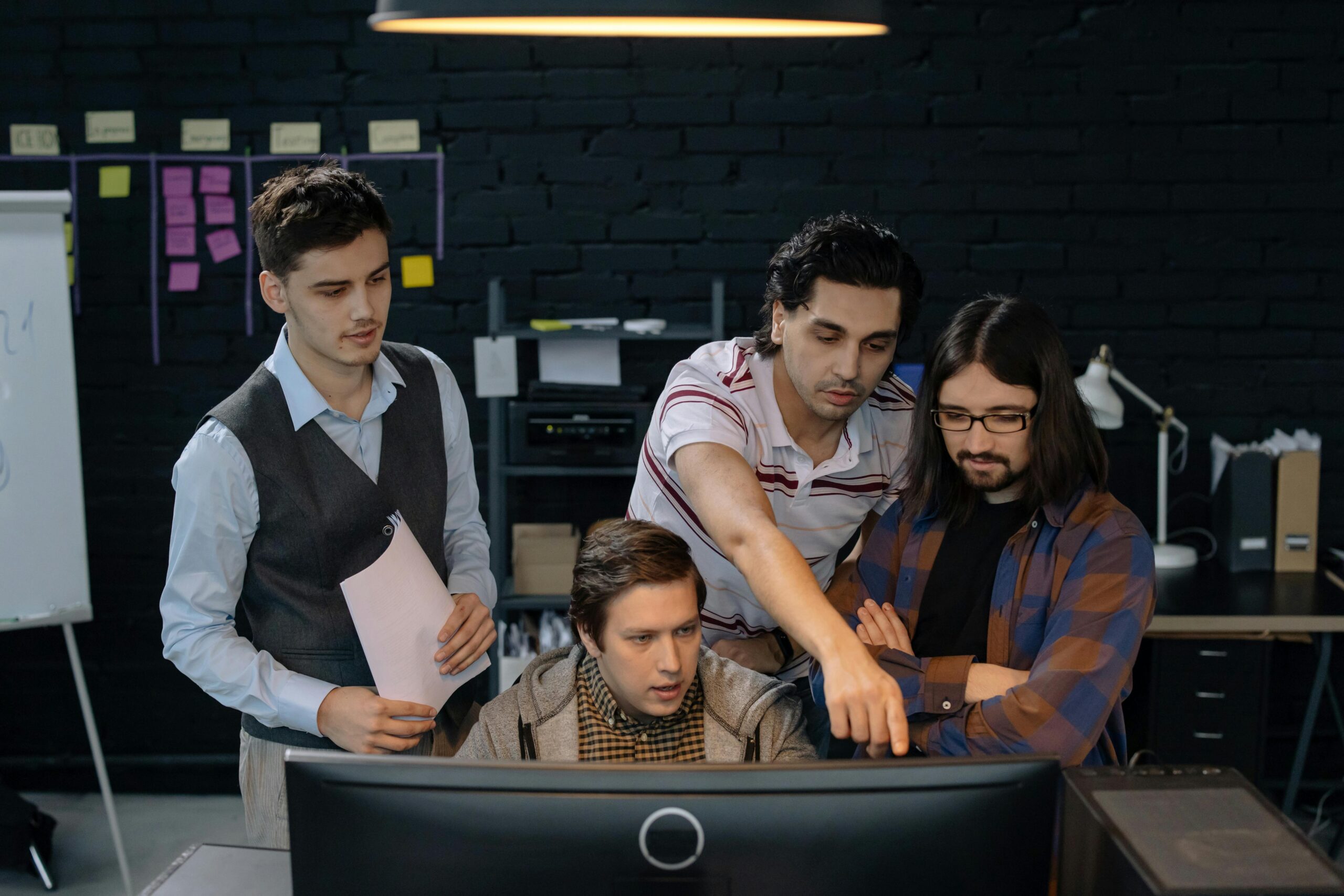 Four focused men collaborating over a computer in a modern office setting with a black brick wall and sticky notes in the background.