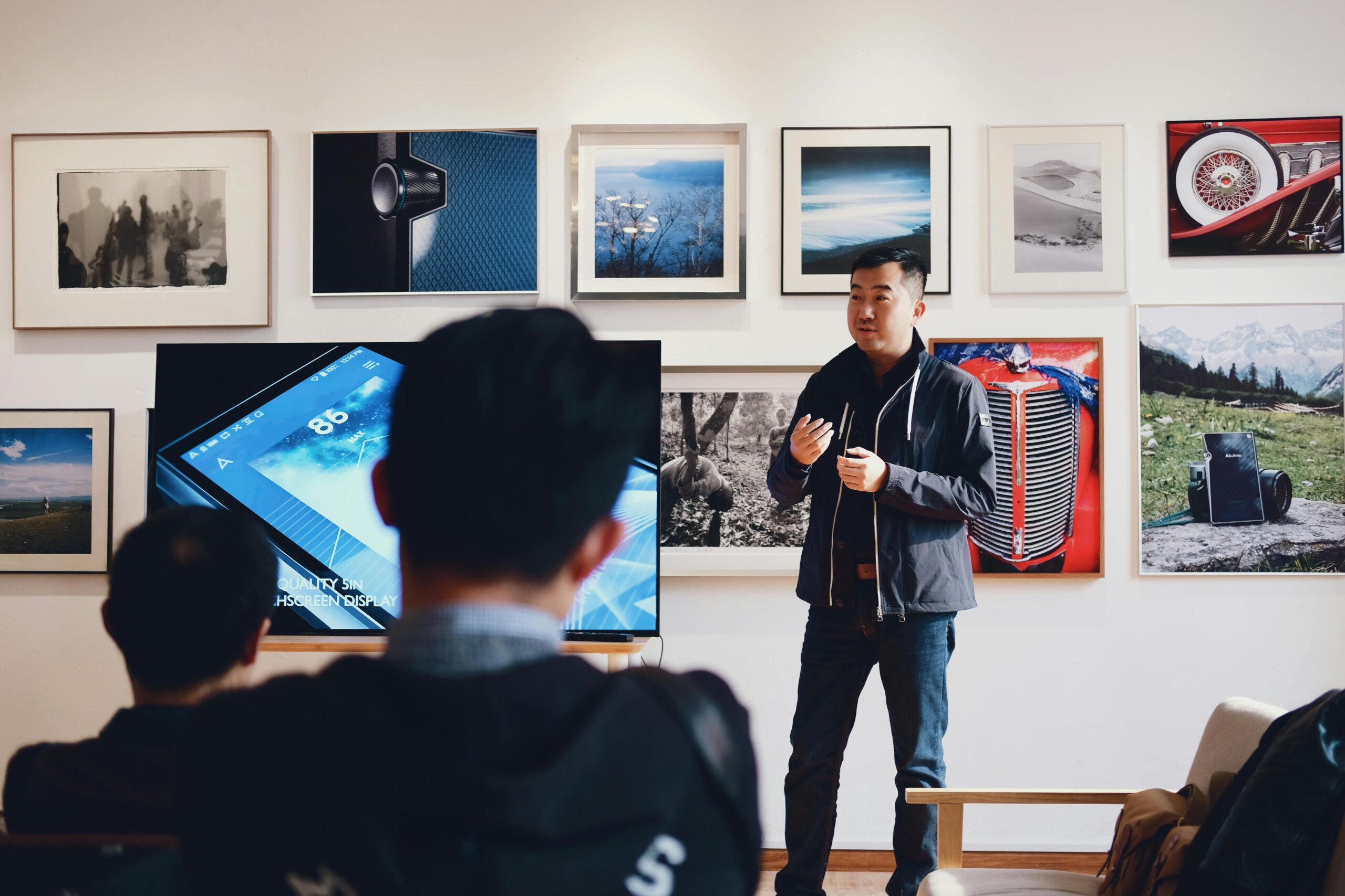 A presenter speaking in front of a screen with visual aids, surrounded by various framed photographs on the wall.
