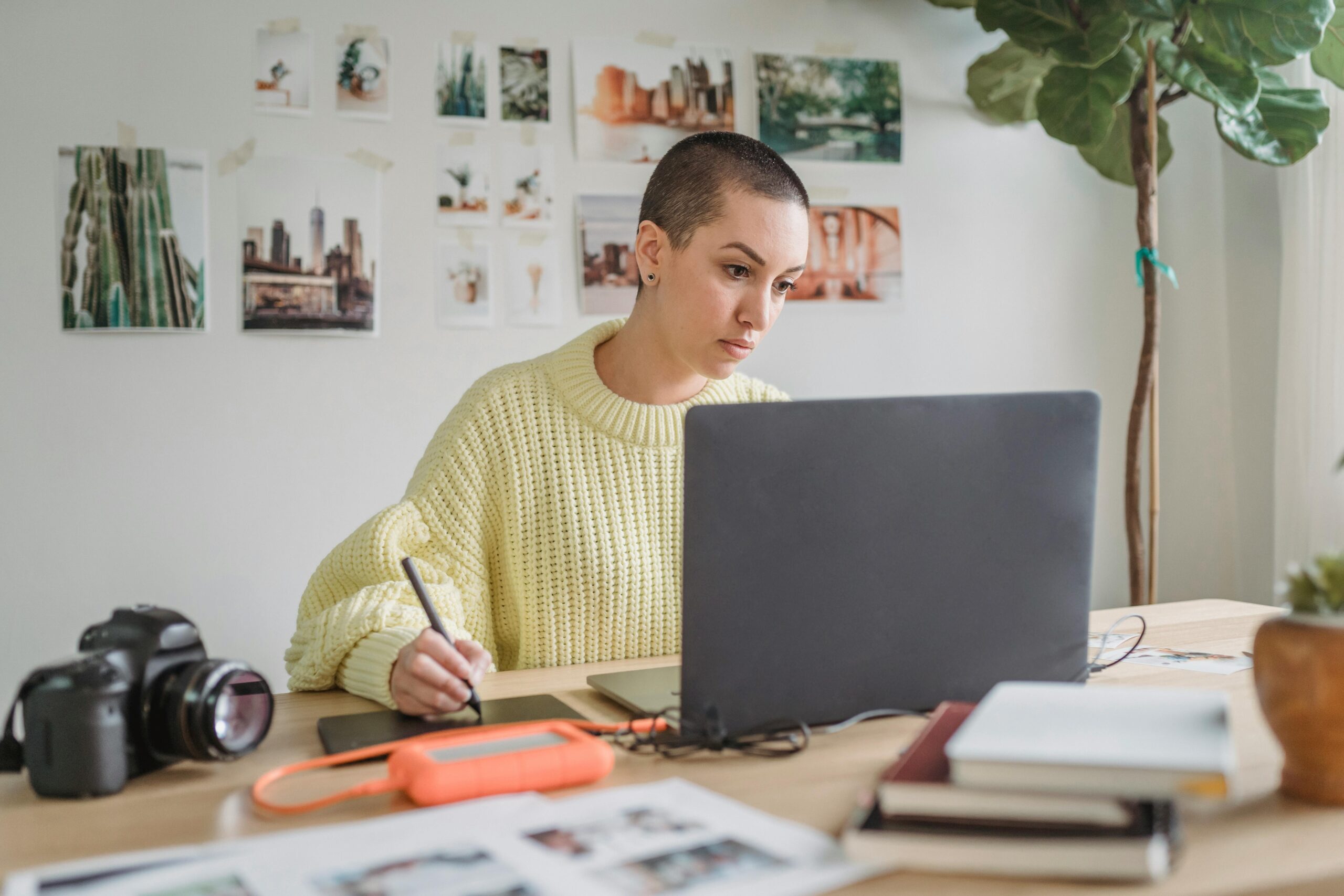 Focused individual working at a desk with a digital tablet, laptop, and cam-velascoera, against a backdrop of photo prints on the wall.