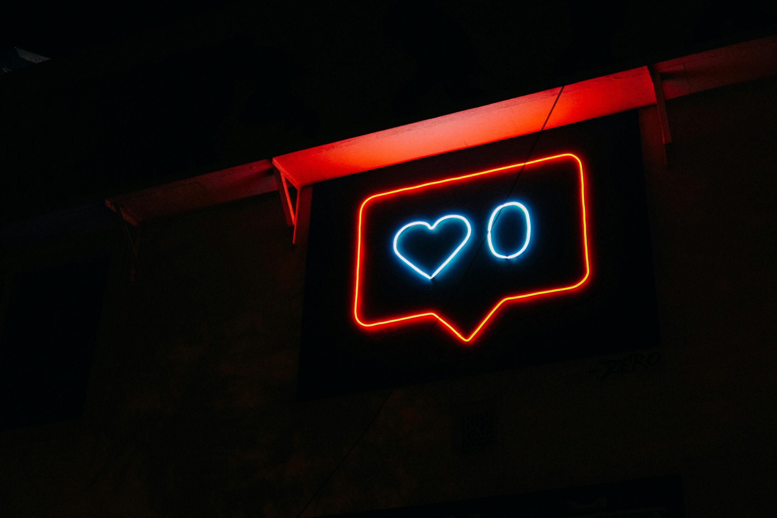 A neon sign of a message bubble with a heart and a zero, glowing in blue and red, against a dark background.