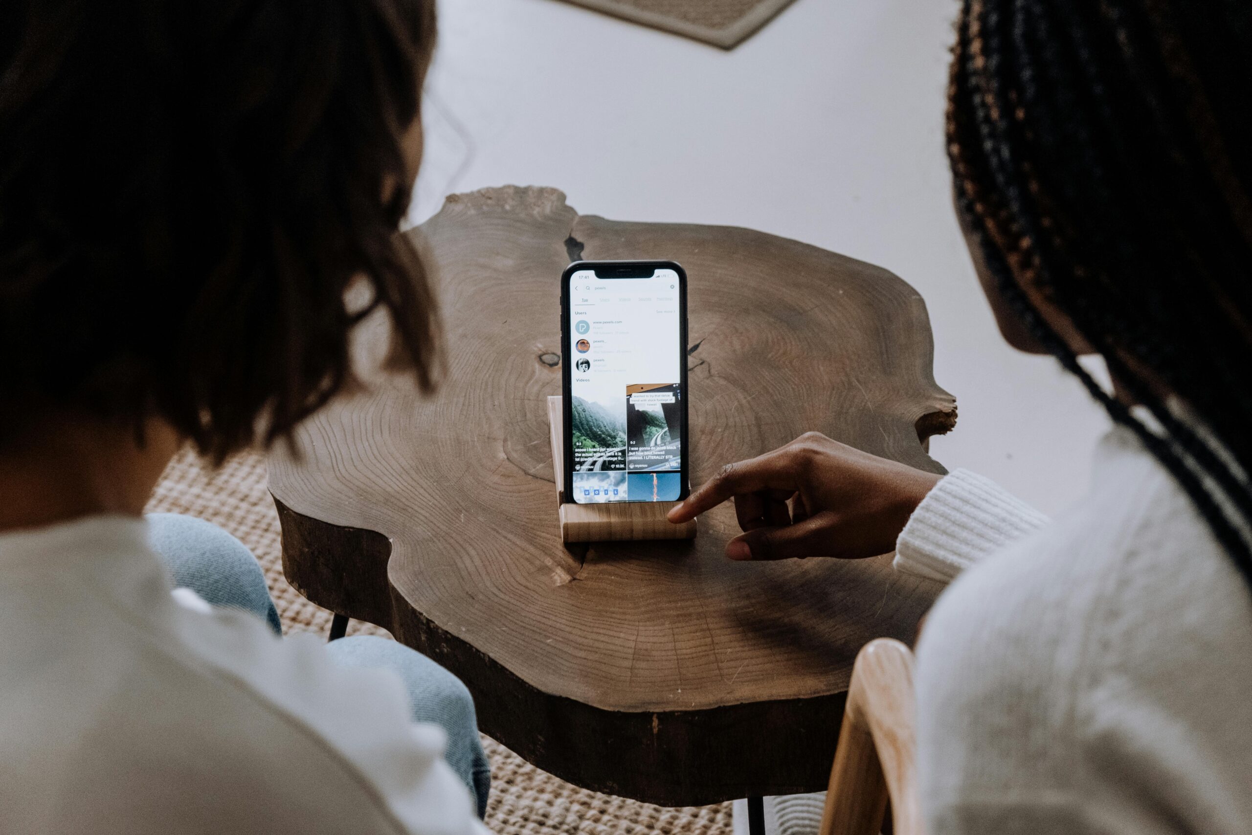 Two people seated at a wooden table viewing a smartphone propped up on a stand.
