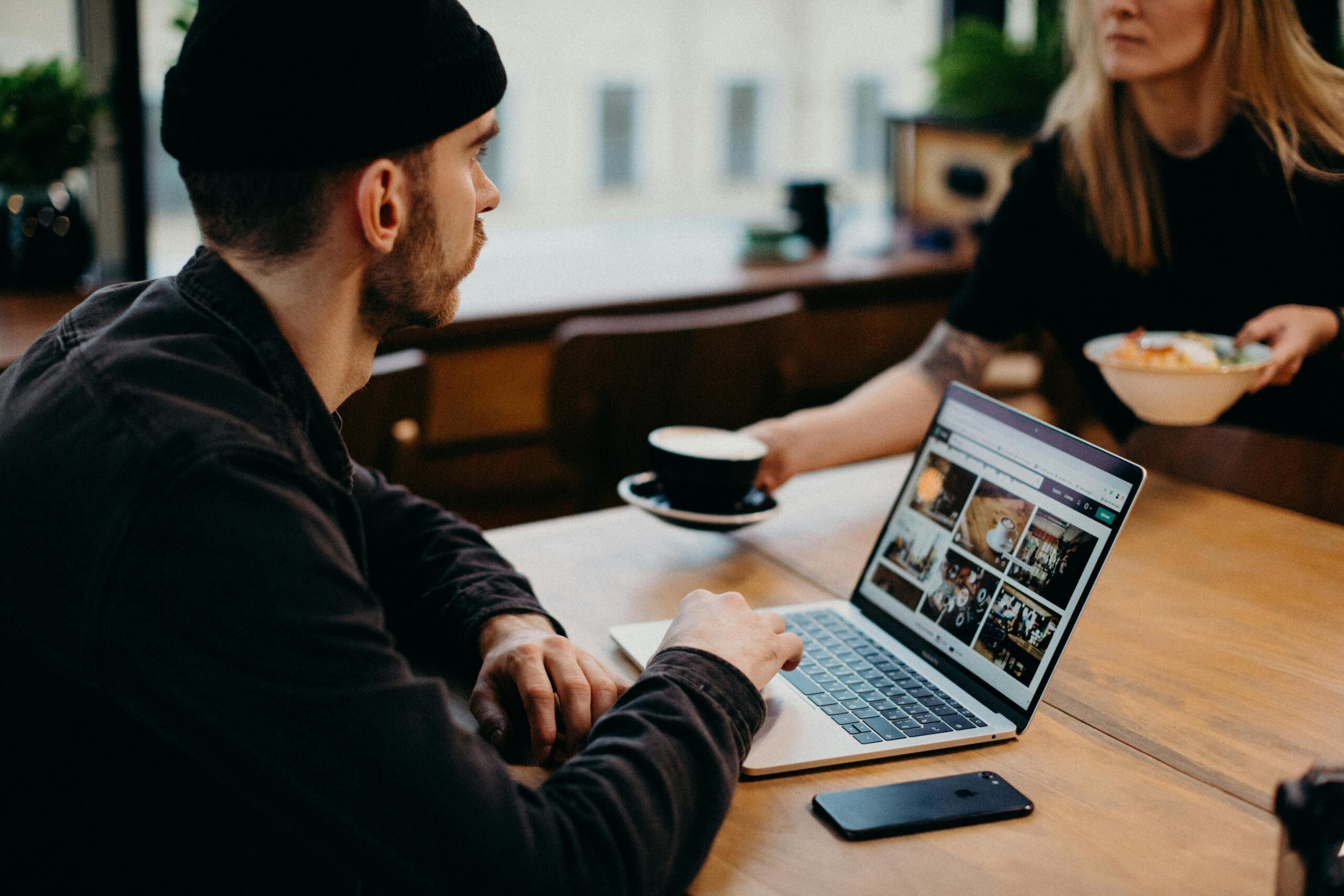 A person in a beanie sitting at a table with a laptop showing a photo gallery, while another person with a plate of food is across the table.