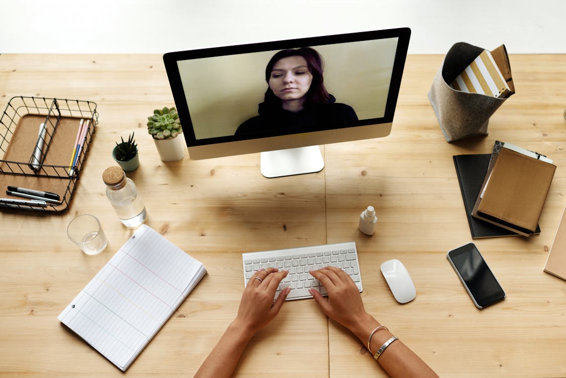 Over-the-shoulder view of a person typing on a keyboard, video calling with a woman on the computer screen.