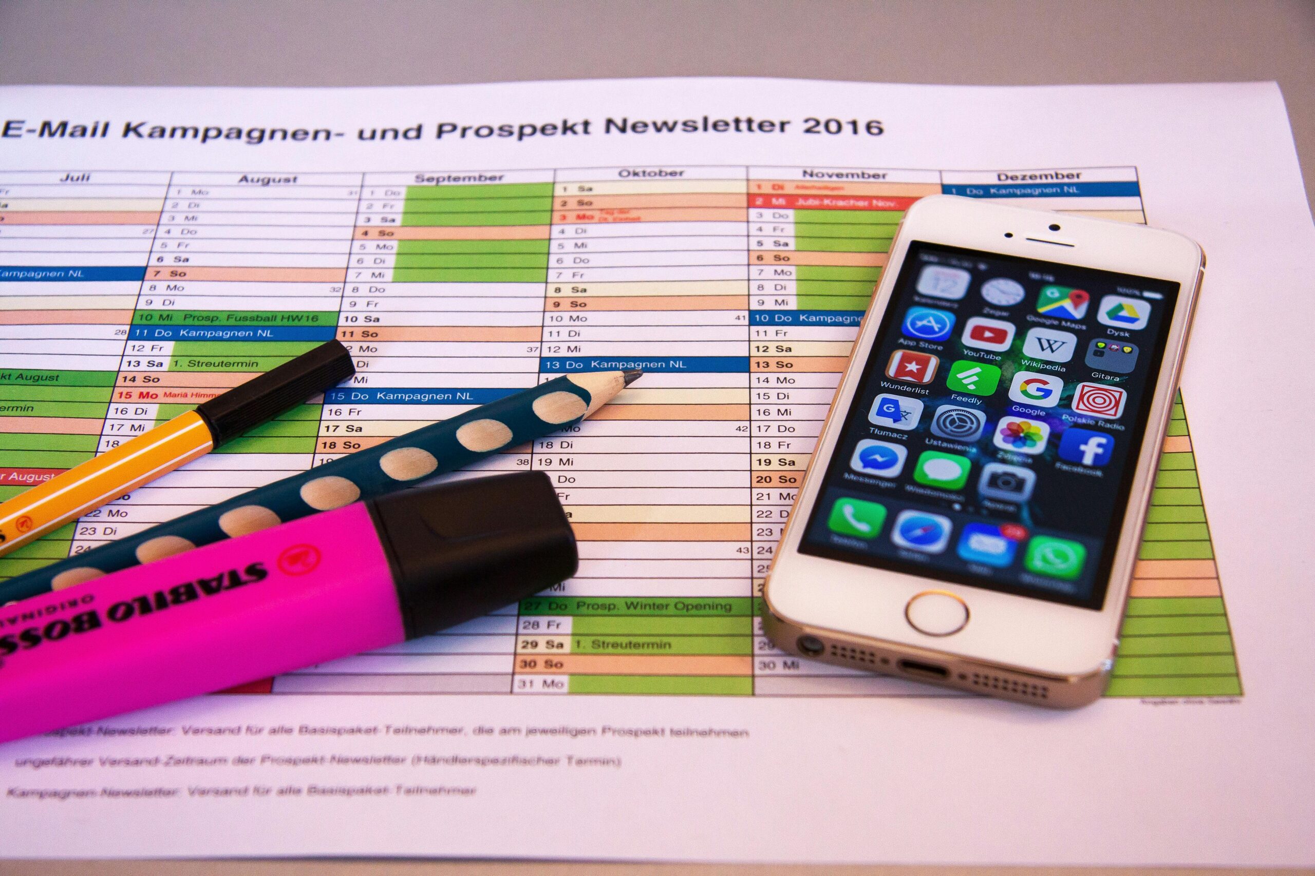 A printed marketing cam-velascopaign and newsletter schedule for the months of July to December 2016, with a smartphone, pencils, and a highlighter pen on top.
