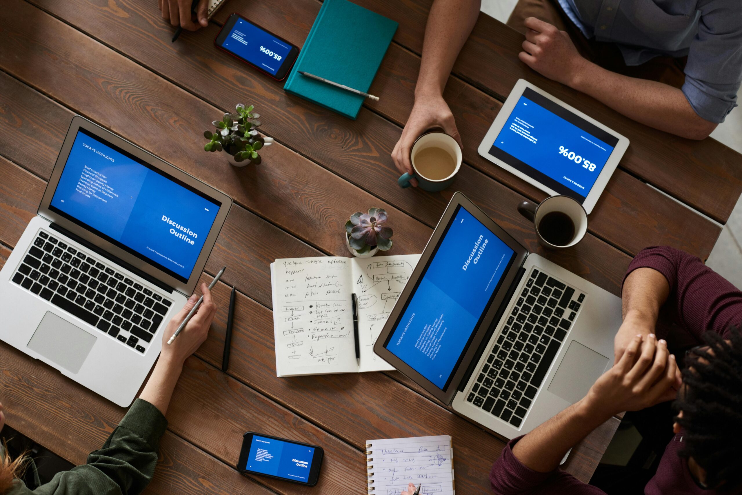 A team of professionals engaged in a collaborative work session around a wooden table with various digital devices displaying a cohesive blue-themed presentation.