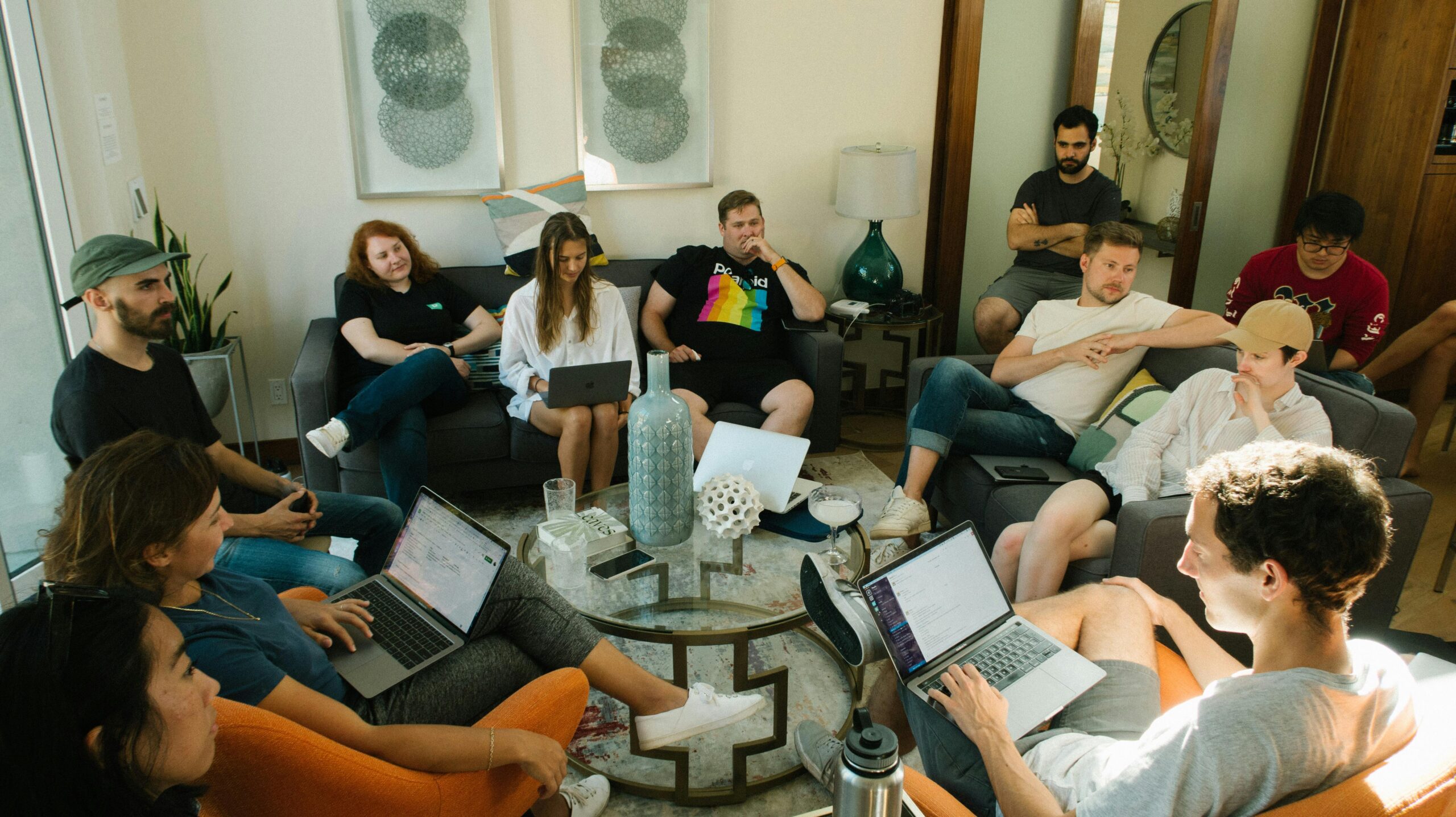 A diverse group of people sitting in a casual living room setting, actively participating in a group discussion with laptops in their laps.