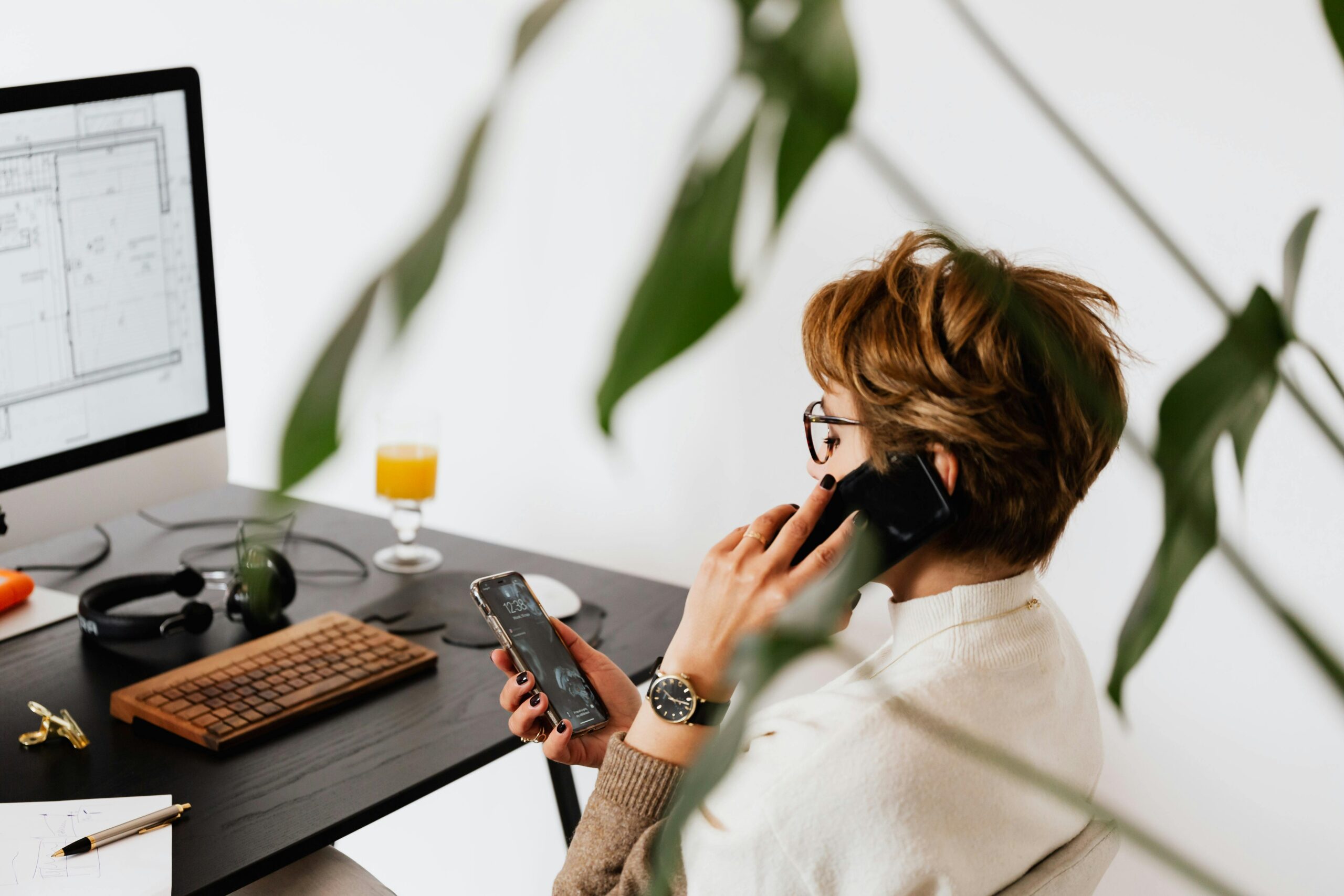 A professional woman in a sweater talking on a black landline phone, with another smartphone in her hand, in a modern office with a computer, houseplants, and a glass of orange juice on the desk.
