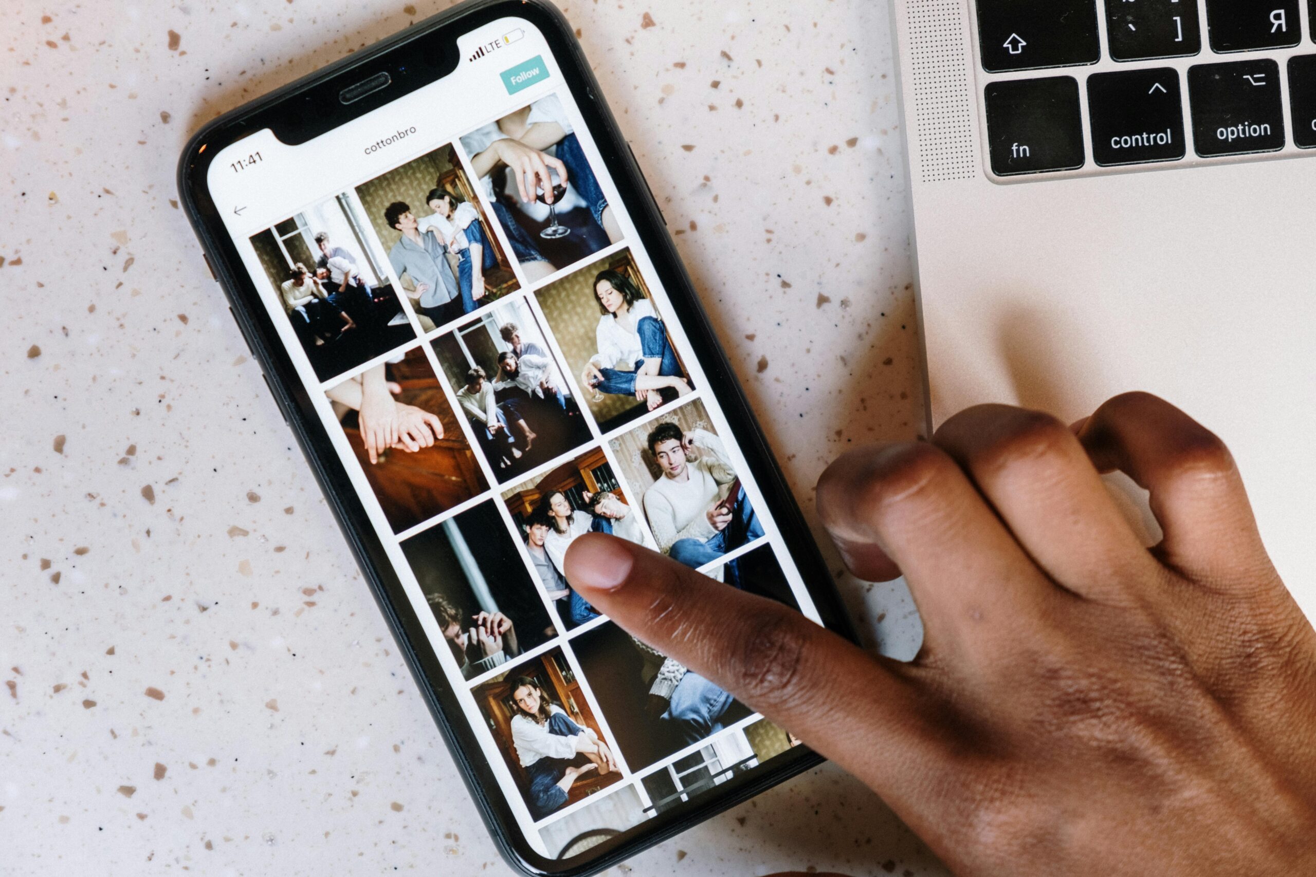 A hand scrolls through a photo gallery on a smartphone, showing a grid of images with various people, alongside a laptop on a speckled surface.